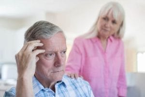 An older man is displaying signs of dementia, as an older woman observes him attentively.