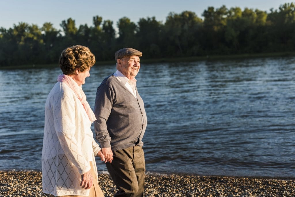 An older couple walking along the shore of a river.