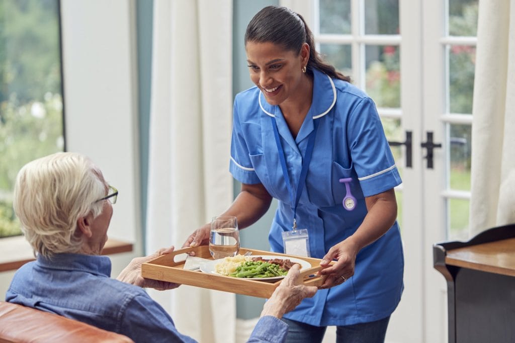 Female Care Worker In Uniform Bringing Meal On Tray To Senior Man Sitting In Lounge At Home