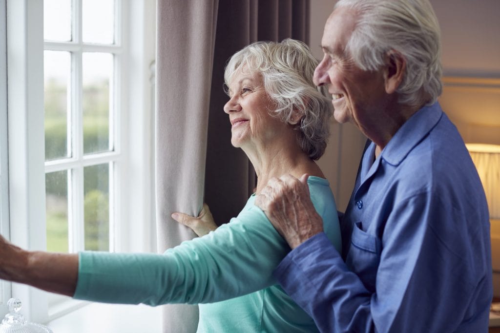 Smiling Senior Couple At Home Wearing Pyjamas Opening Bedroom Curtains And Looking Out Of Window