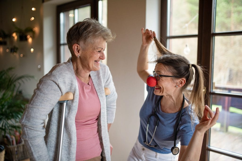 Healthcare worker or caregiver with red nose visiting senior woman indoors at home, having fun