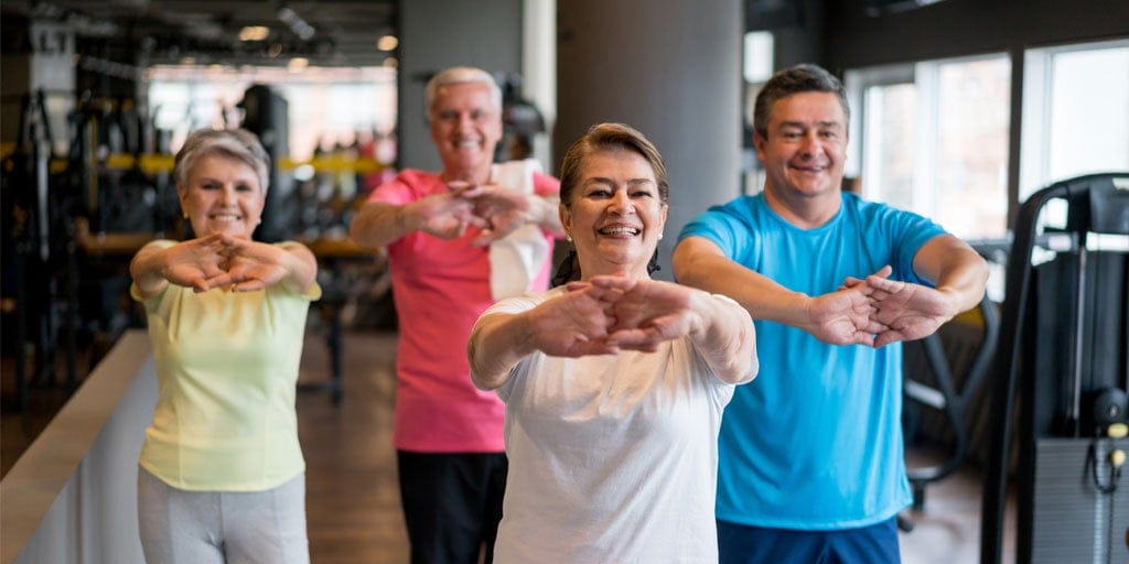 Four older adults are standing in a row and stretching their arms out in front of them inside a gym. They appear to be participating in a fitness class.