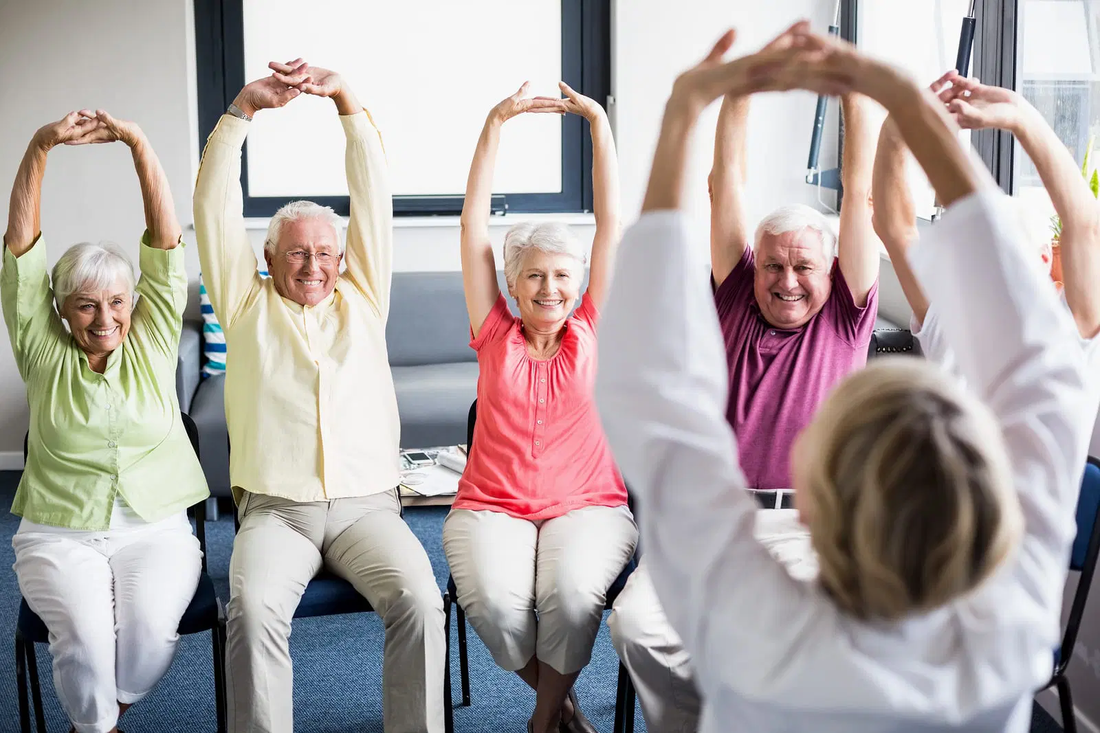 A group of elderly people seated in chairs, led by an instructor, perform synchronized arm stretches in a well-lit room with large windows.