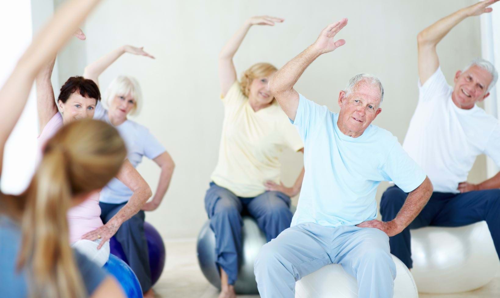 A group of seniors seated on exercise balls, engaging in a group fitness class with their arms raised overhead.