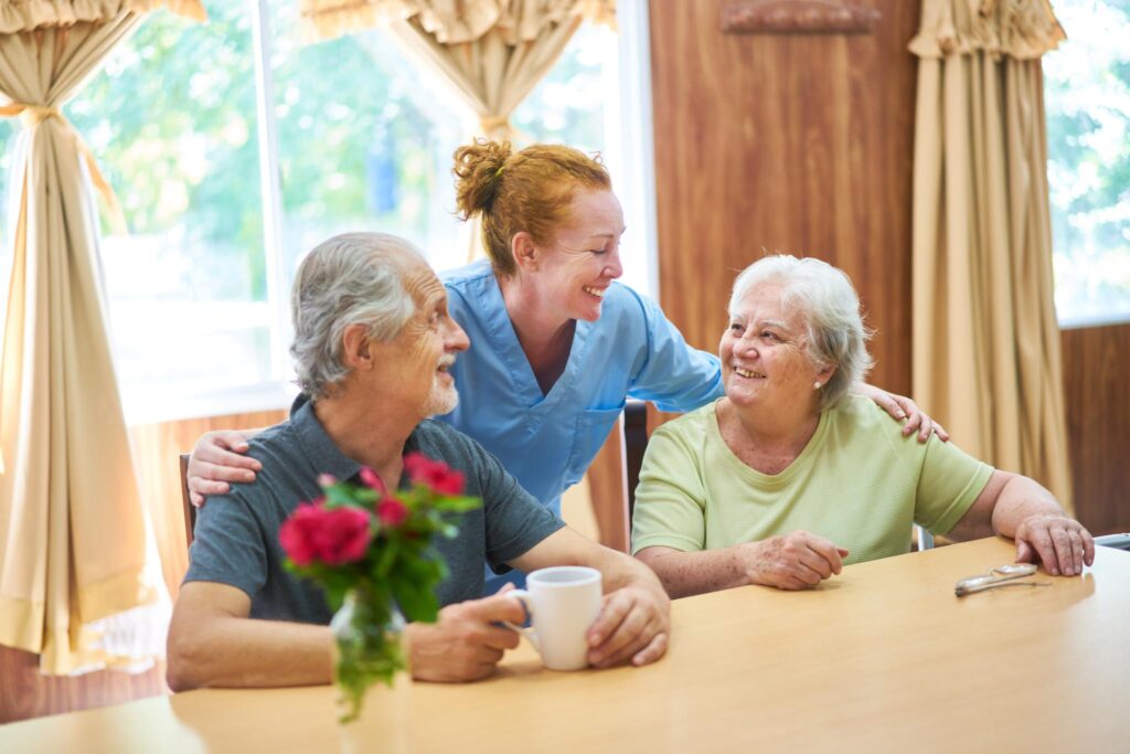 A nurse in blue scrubs smiles and interacts with an elderly man holding a coffee mug and an elderly woman at a table with flowers in a vase, showcasing the warm atmosphere of this adult day care. Curtains and a window in the background add to the inviting setting.