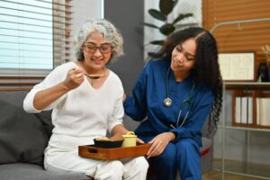 An older woman in a white outfit eats soup while a healthcare professional in blue scrubs with a stethoscope provides assistance. They are indoors, sitting on a couch.