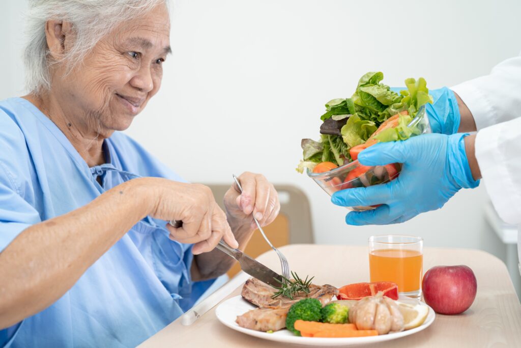 Elderly person in a hospital gown eats a meal featuring vegetables and meat. A gloved hand offers a bowl of salad. A glass of juice and an apple are on the table.