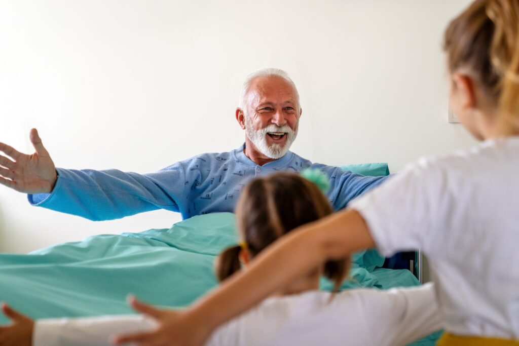 Older man in a hospital bed with arms open wide, smiling at two children reaching towards him.