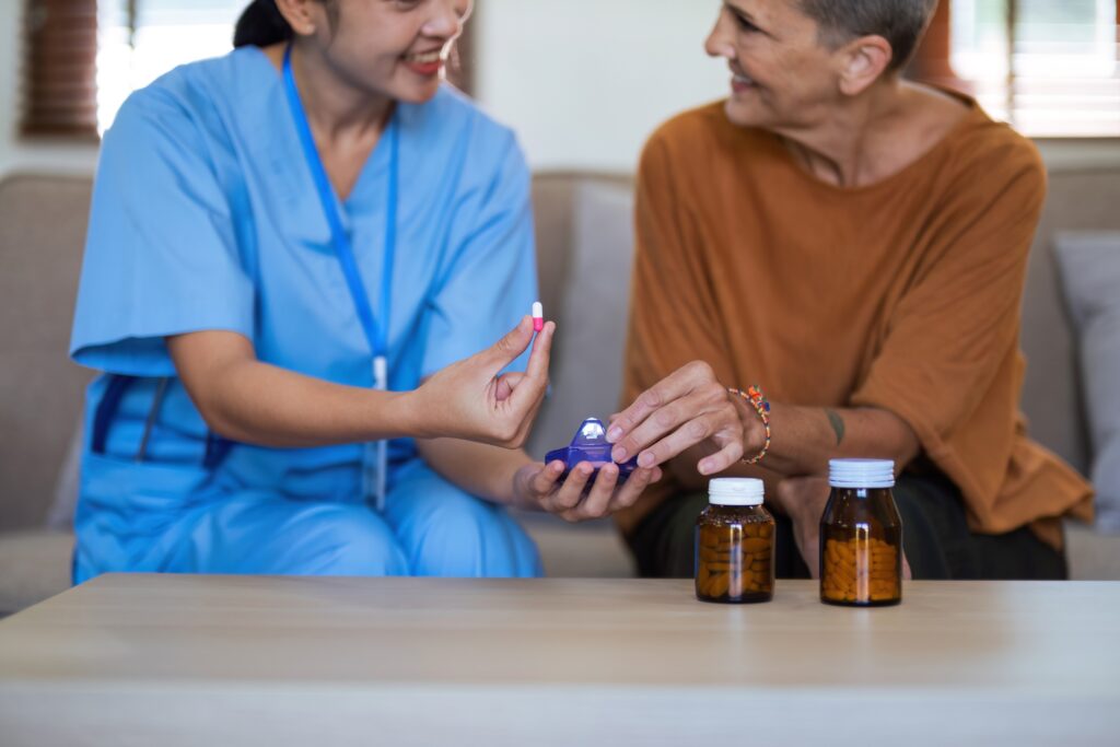 A healthcare worker in blue scrubs gives a pill to a seated person. Two medicine bottles are on the table in front of them.