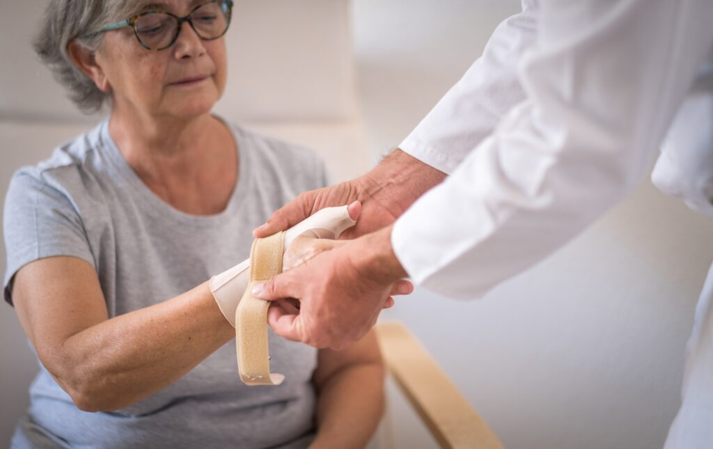 A doctor wraps an older woman's injured hand with a bandage. She is sitting and looking at her hand.