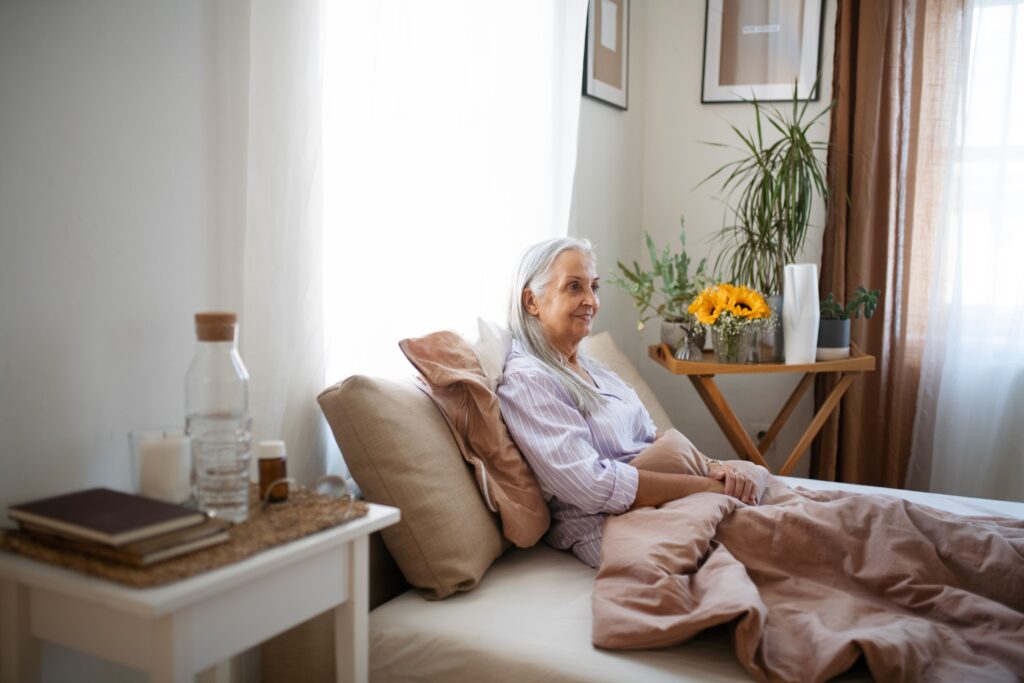 Elderly woman with gray hair sits on a bed, covered with a blanket, in a well-lit room with plants and decor.