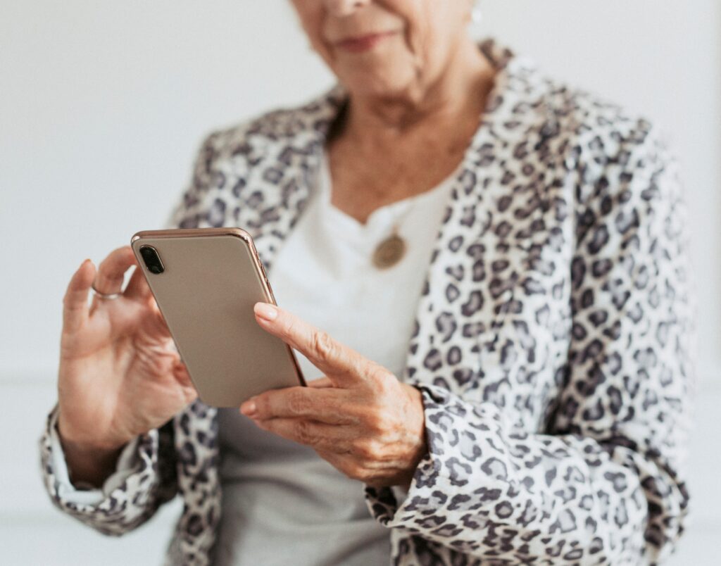 An elderly woman in a leopard print jacket uses a smartphone, focusing on the screen with both hands.