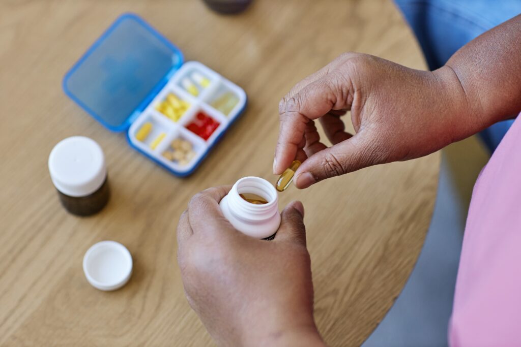 Person holding a pill from a white bottle, with a pill organizer containing various pills on a wooden table.