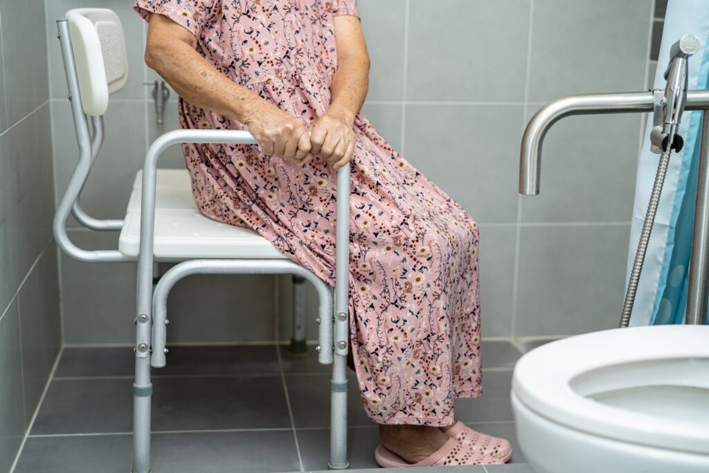 Elderly person sitting on a shower chair in a bathroom with grab bars, wearing a patterned pink dress and slippers.