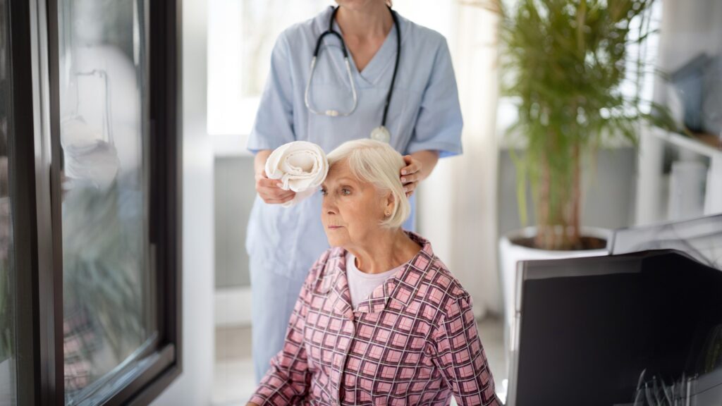 A nurse in scrubs holds a towel behind an elderly woman seated in a wheelchair. The woman wears a checkered shirt. A plant is visible in the background.