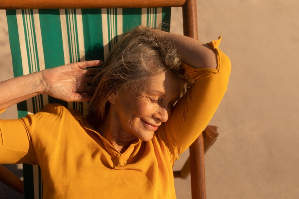 A woman in a yellow shirt relaxes in a striped deck chair on a sandy beach, smiling with her eyes closed, and one arm behind her head.