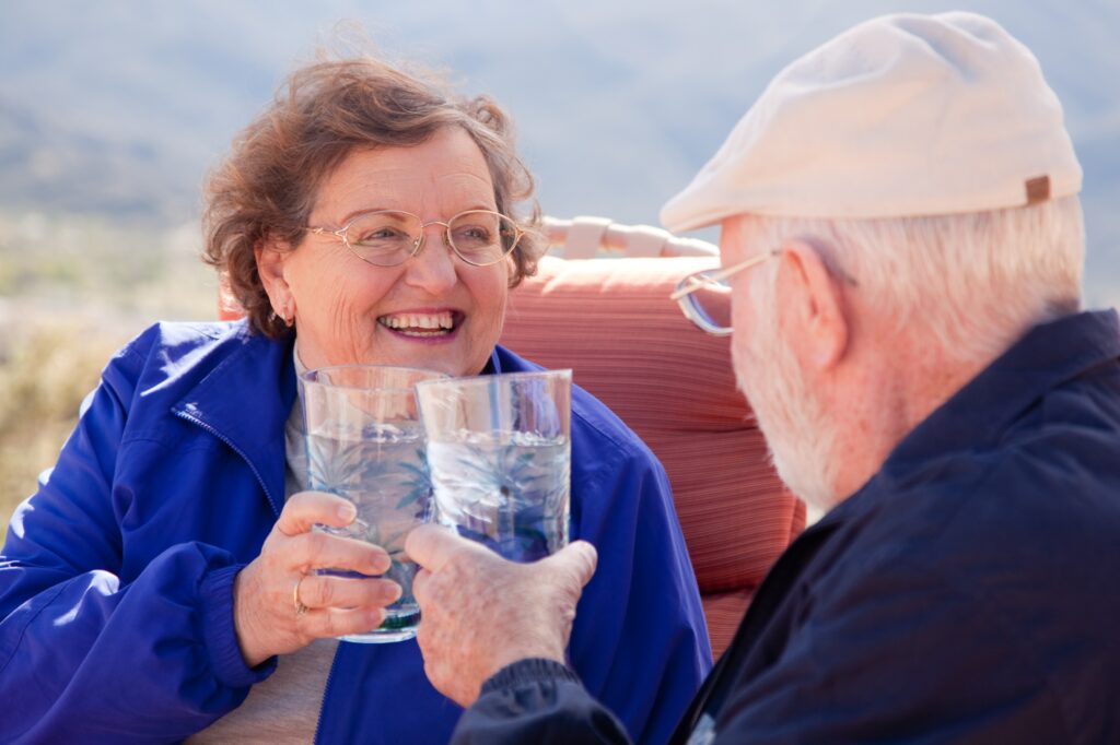 Two elderly people cheerfully clink glasses of water while sitting outside on a sunny day.