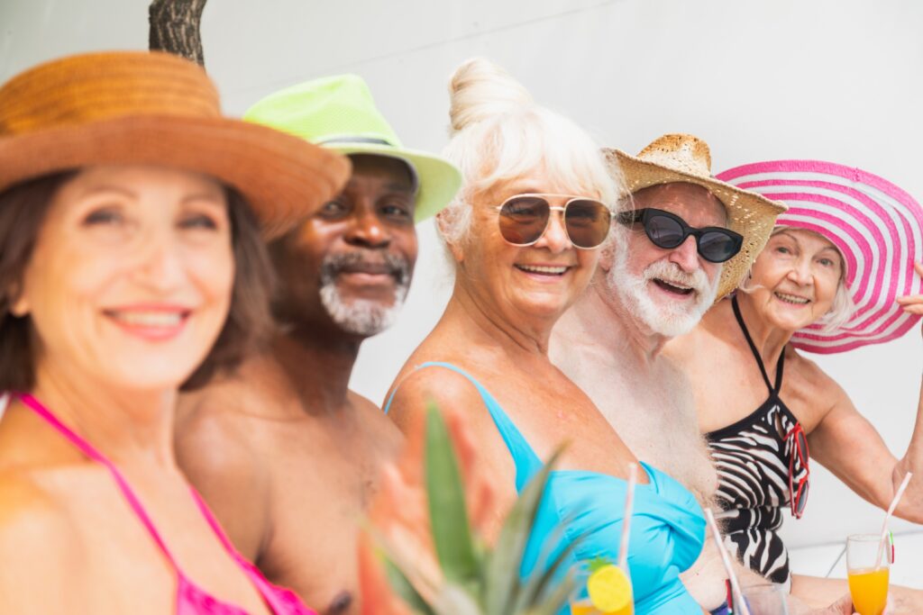 Five elderly people wearing hats and swimsuits smile while sitting outdoors, holding drinks.