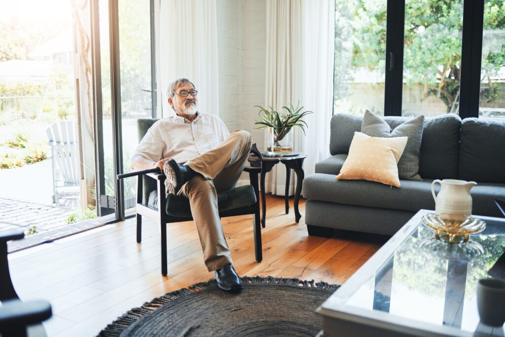 An older man sits relaxed in a modern living room, with one leg crossed over the other. The room features large windows, light curtains, a gray sofa, and a round rug on a wooden floor.