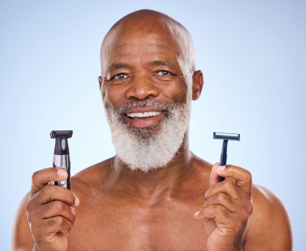 A man with a beard holds an electric razor in one hand and a manual razor in the other, smiling against a blue background.