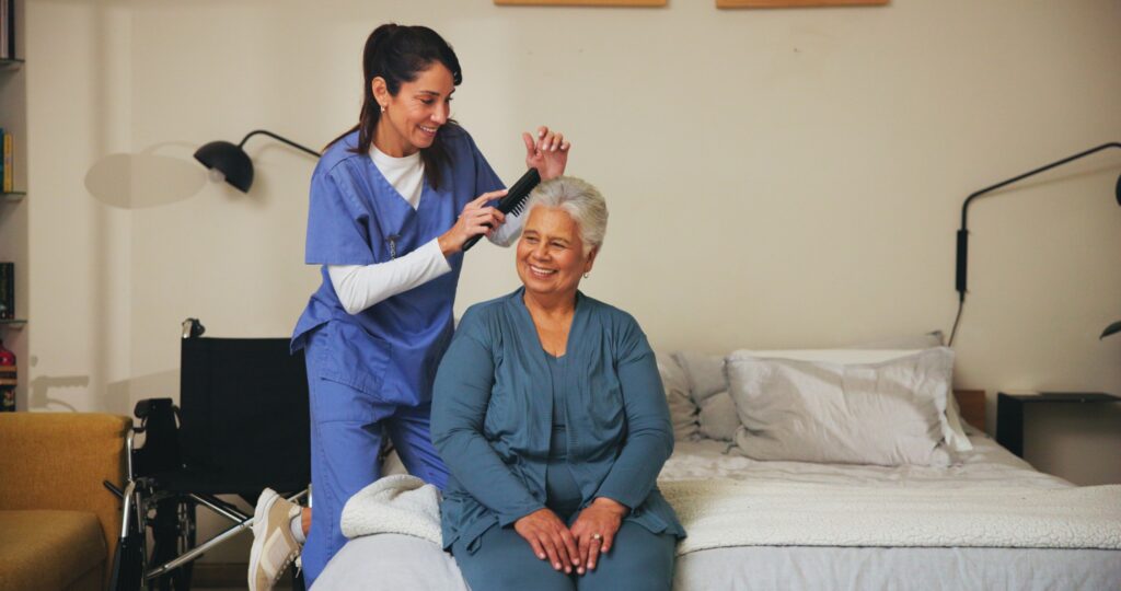 A caregiver in blue scrubs brushes the hair of a smiling elderly woman sitting on a bed in a cozy room.