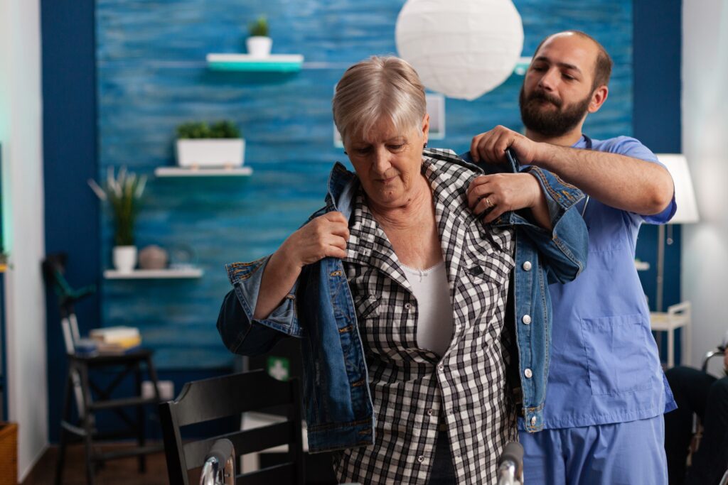 A caregiver assists an elderly woman in wearing a denim jacket in a room with blue walls and shelves with plants.