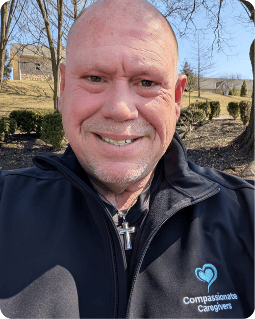 A person in a dark jacket with "Compassionate Caregivers" logo and heart design smiles outdoors, wearing a cross necklace. There are trees and houses in the background.