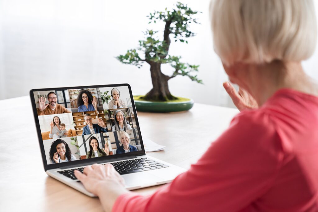 A person in a red shirt participates in a group video call on a laptop. A small bonsai tree is visible on the desk.