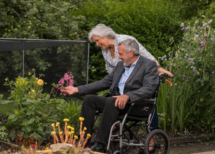 An older adult woman and man in a wheelchair examine flowers in a garden. The woman leans over, smiling, while the man reaches out to touch a plant.