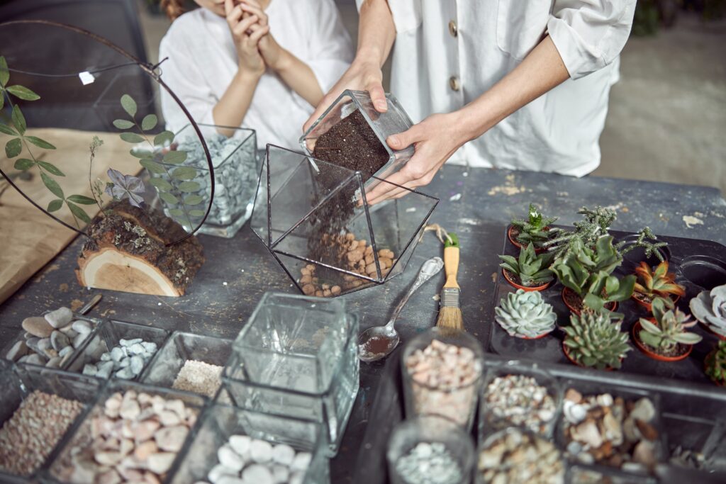 Person pouring soil into a geometric glass container on a table with various plants, stones, and gardening tools.