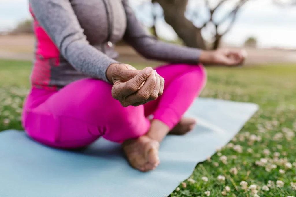 Person in pink leggings and a gray top sits cross-legged on a yoga mat outdoors, meditating with hands on knees.