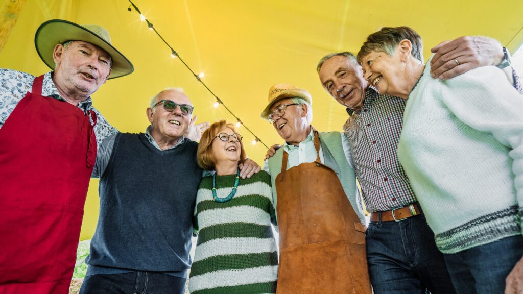 A group of six older adults smiling and standing under a yellow tent. Some wear aprons and hats. They have their arms around each other.