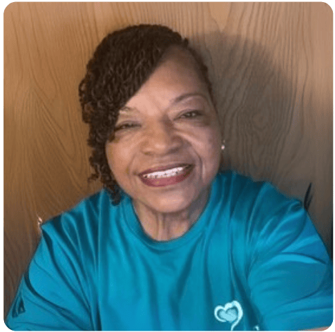 A woman with short, curly hair smiles at the camera. She is wearing a teal shirt with a small heart design on the chest and is posed in front of a wooden background.