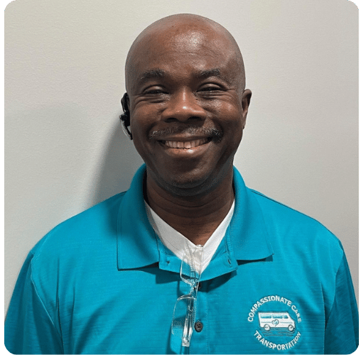 Smiling man wearing a teal polo shirt with a "Compassionate Care Transportation" logo, glasses hanging from his collar, standing against a plain light-colored wall.