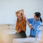 A healthcare professional assists an older woman with a seated arm stretch in a well-lit room during a physical therapy session.
