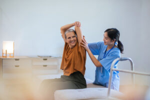 A healthcare professional assists an older woman with a seated arm stretch in a well-lit room during a physical therapy session.