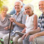 Four older adults sit outdoors on a bench, smiling at the camera. One holds a walking cane, another holds an open book. The background is green and sunlit.