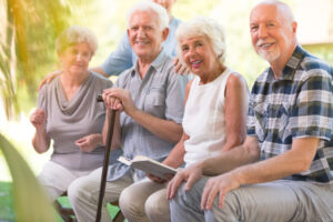 Four older adults sit outdoors on a bench, smiling at the camera. One holds a walking cane, another holds an open book. The background is green and sunlit.