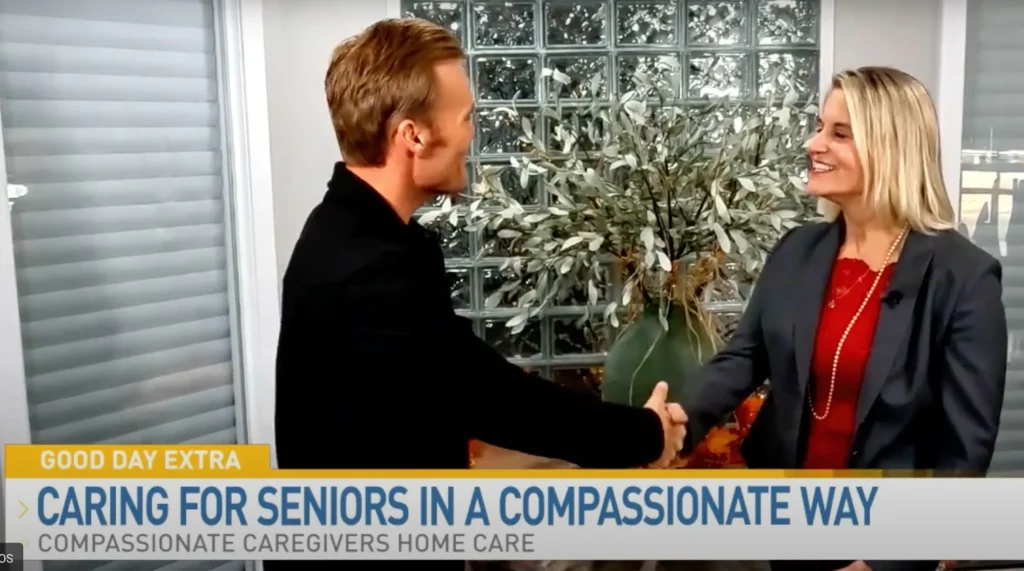 Two people shake hands and smile during a TV interview segment about compassionate home care for seniors, with a plant and glass block wall in the background.