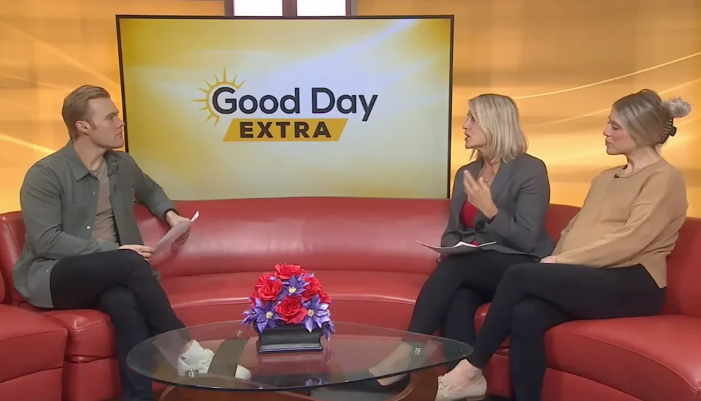 Three people sit on red couches in a TV studio with a "Good Day Extra" sign displayed behind them, discussing home care. A glass coffee table with a flower arrangement is in the foreground.