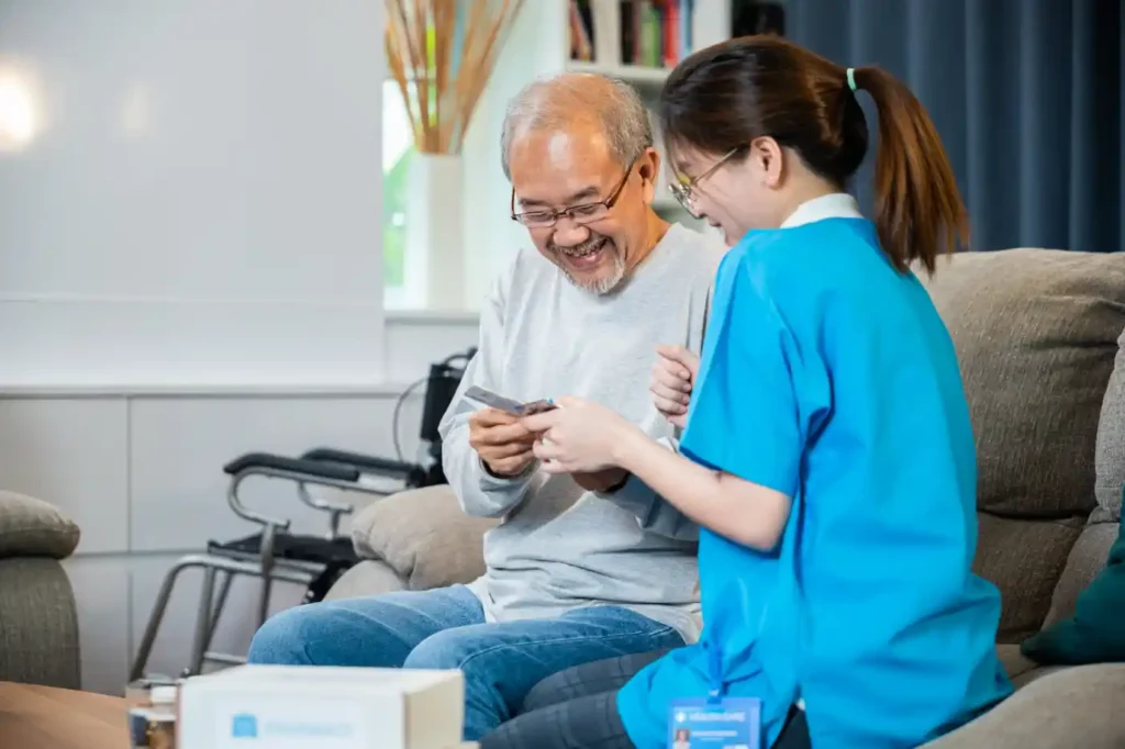 An elderly man sits on a couch smiling and looking at a card, while a healthcare worker in blue uniform—providing home care services—sits beside him. A wheelchair is visible in the background.