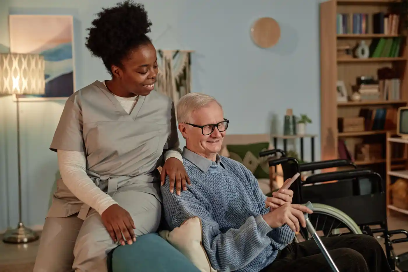 A caregiver in scrubs smiles while talking to an older man in glasses seated next to a wheelchair, reflecting the warmth of assisted living in Columbus, Ohio.