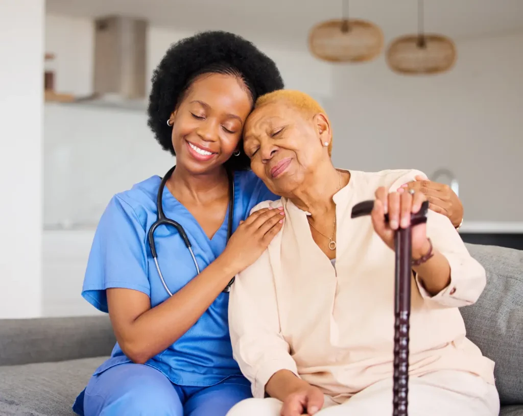 A nurse in blue scrubs sits on a couch beside an elderly woman with a cane, both smiling and leaning their heads together, reflecting the warmth and compassion of quality home care in Columbus.