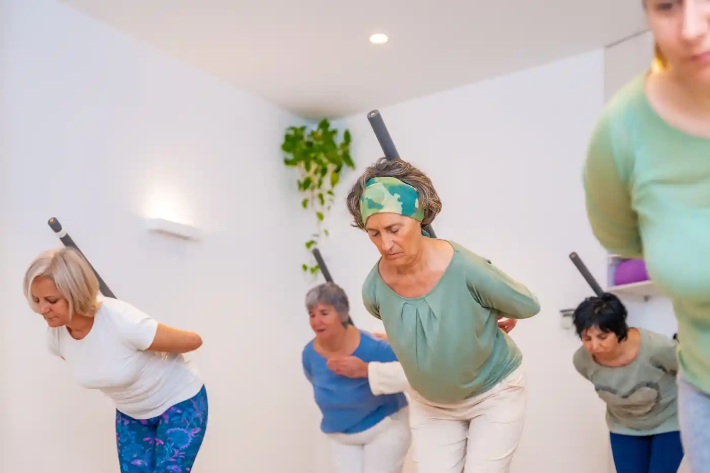 Five women participate in a fitness class, bending forward with sticks across their shoulders in a bright, minimalist room—an example of the healthy lifestyle supported by Upper Arlington Home Care.