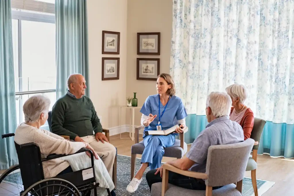 A healthcare worker in blue scrubs leads a discussion with four elderly people, one in a wheelchair, highlighting the benefits of home care services in a bright, comfortable room.