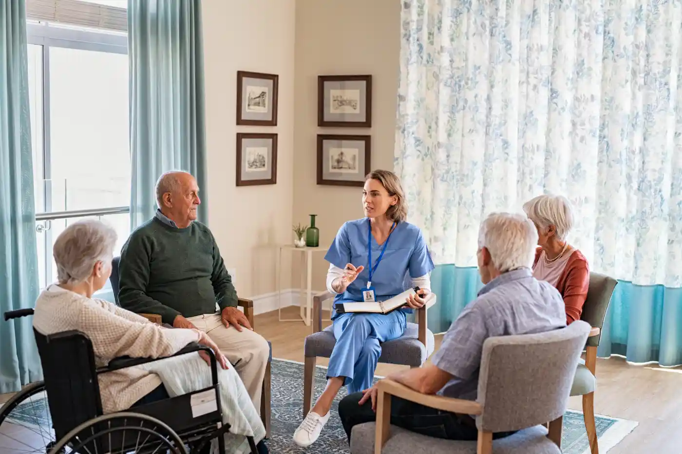 A healthcare worker in blue scrubs leads a discussion with four elderly people, one in a wheelchair, highlighting the benefits of home care services in a bright, comfortable room.