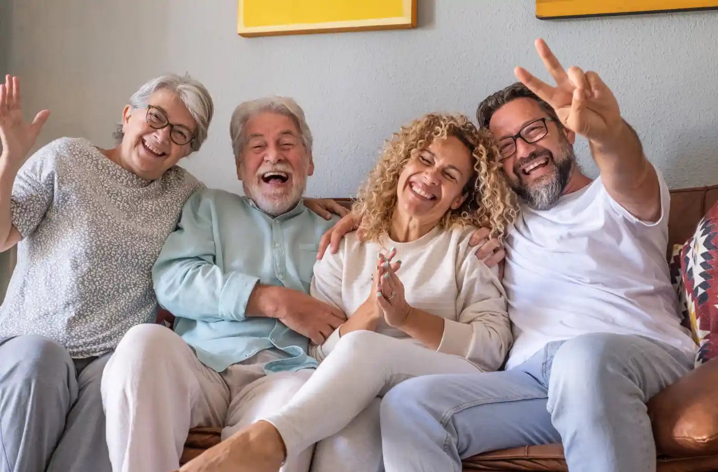 Four adults sit close together on a couch, smiling and laughing. One person holds up a peace sign. They appear to be enjoying each other's company in a casual, indoor setting.
