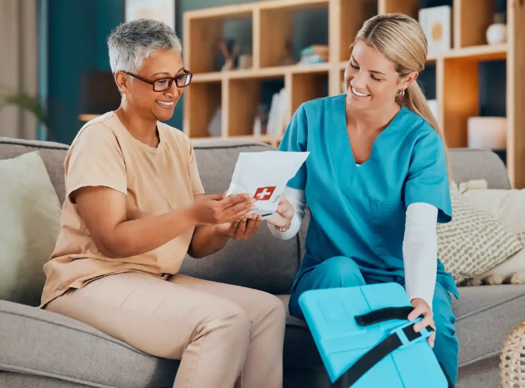 A nurse in blue scrubs hands a medical packet to a smiling woman sitting on a couch in a living room.