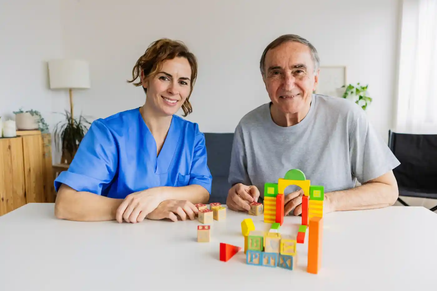 A woman in scrubs and an older man sit at a table smiling, building a structure with colorful wooden blocks—a warm moment highlighting compassionate caregivers in Columbus senior care.