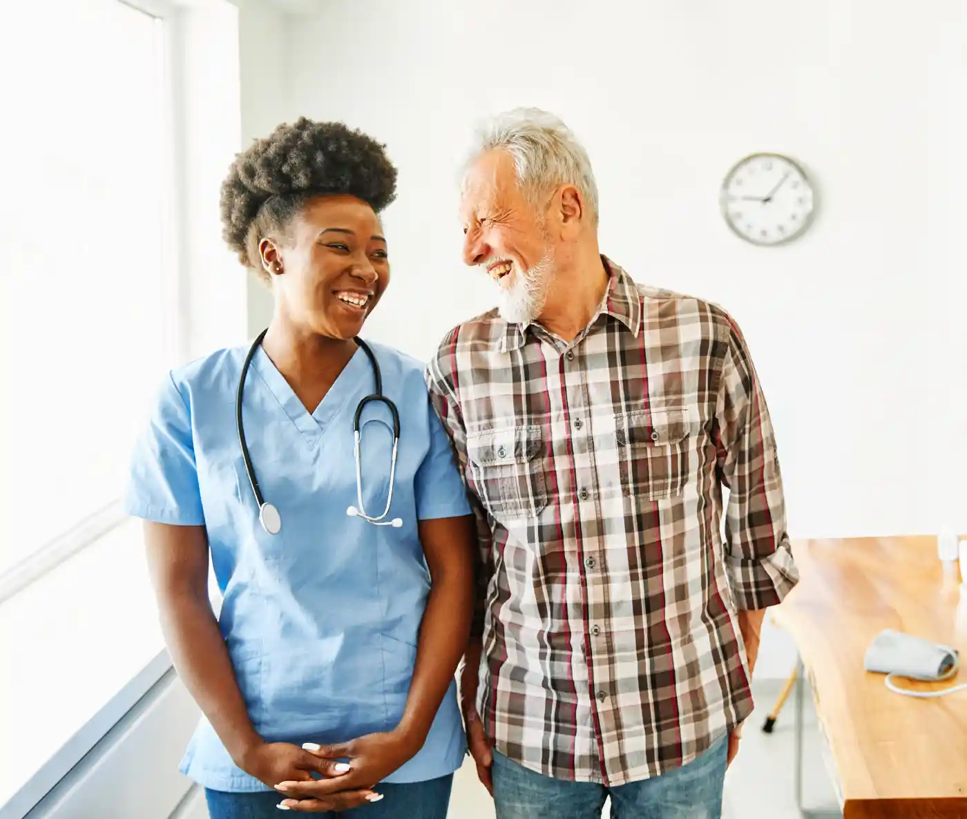 A nurse in blue scrubs stands next to an older man in a plaid shirt, both smiling in a bright room with a wall clock—capturing the warmth of Non Medical Home Care.
